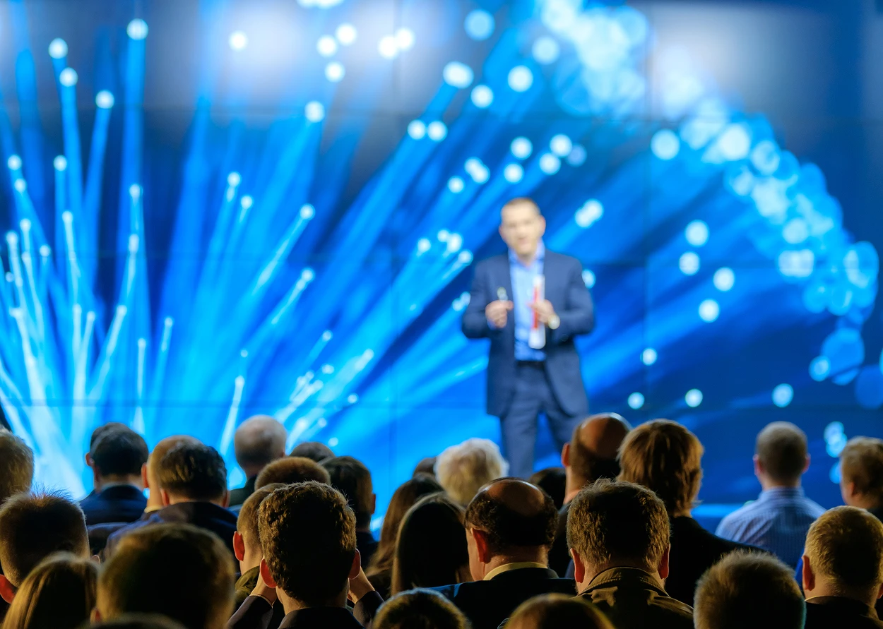 Audience facing a speaker in a suit presenting in front of a large blue abstract background on a screen.