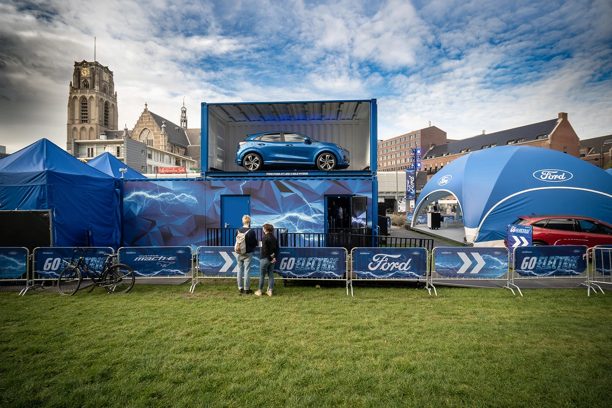 Outdoor Ford display with a blue electric car on an elevated platform, Ford tents, and two people standing by barriers with Ford branding.