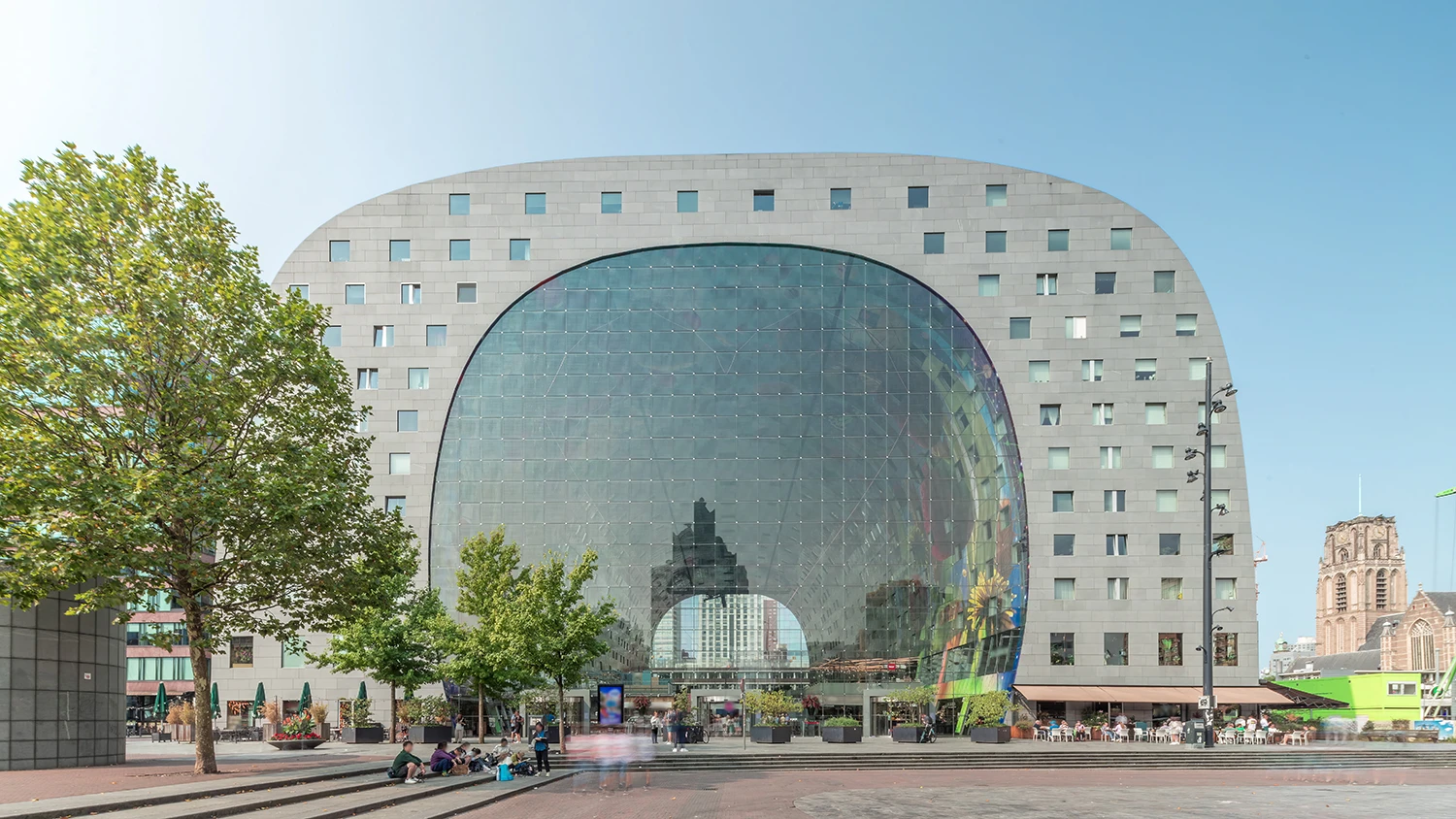 Large modern market hall with a curved glass facade reflecting city buildings and people sitting near trees in the foreground.