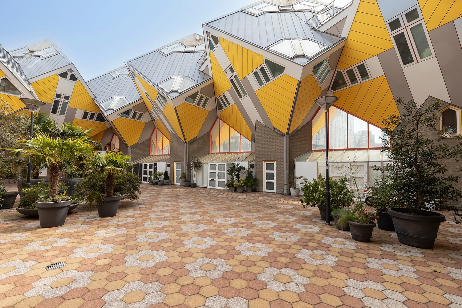Exterior view of yellow cube-shaped houses with tilted roofs and a patterned hexagonal pavement courtyard with potted plants.