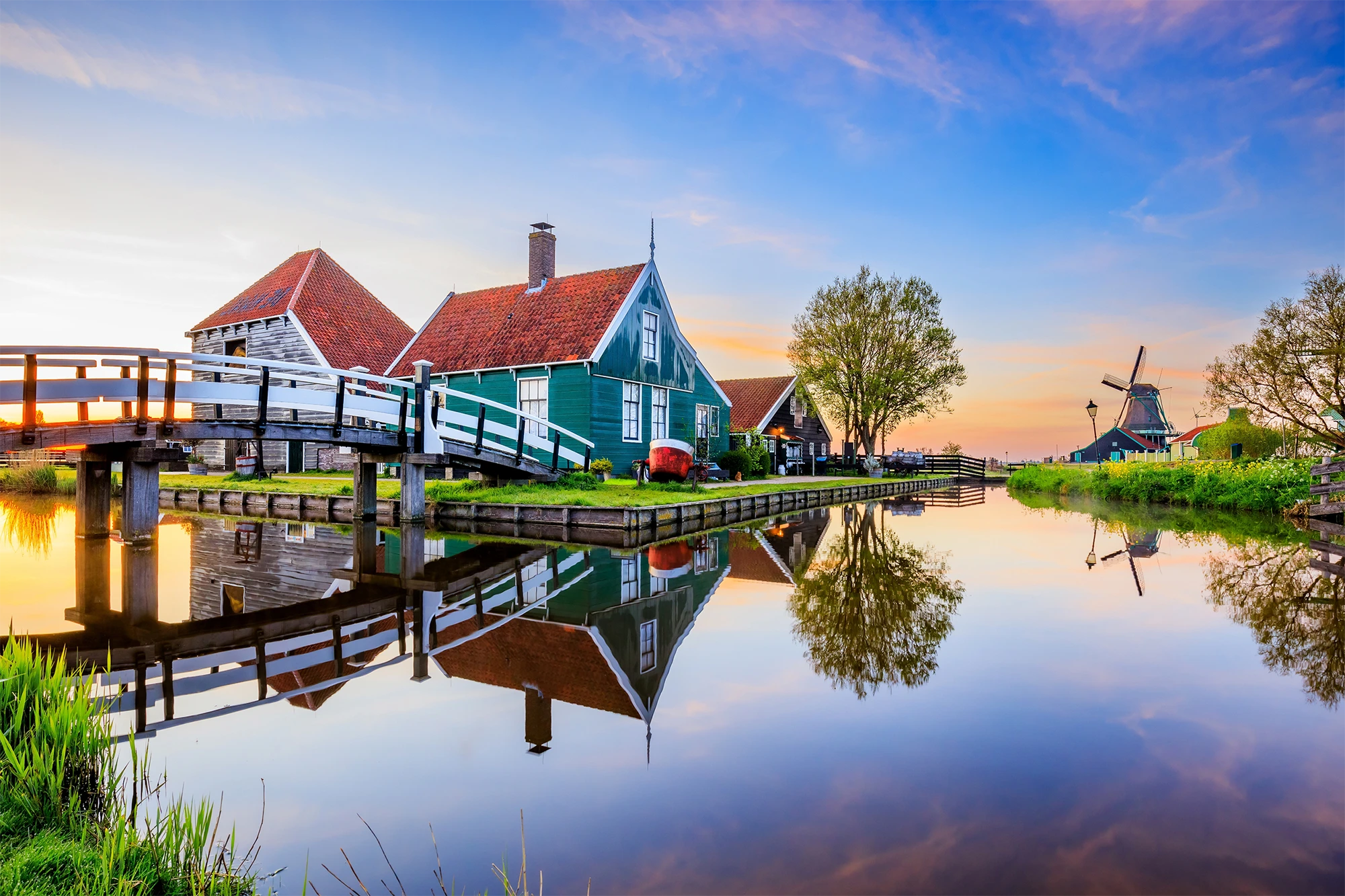 Traditional Dutch houses with red roofs and a white wooden bridge over a calm canal reflecting the buildings and a windmill at sunset.