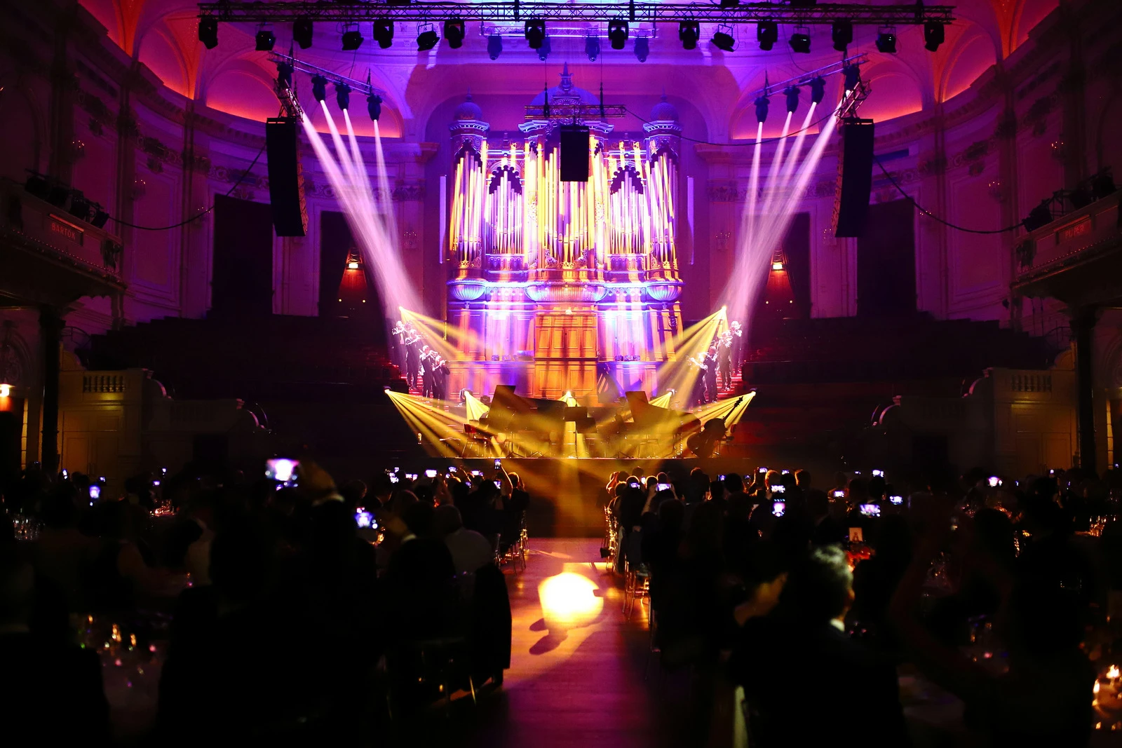 Audience seated in a grand hall watching a stage lit with purple and yellow lights highlighting a large pipe organ and musicians.