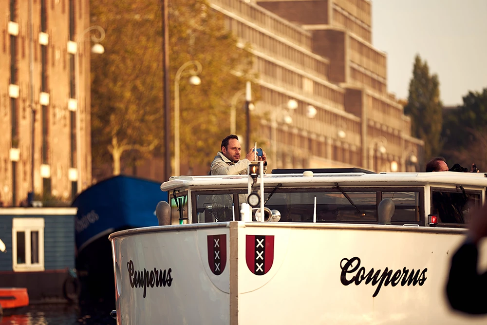 Man taking photo from the upper deck of the boat named Couperus on a canal with buildings and trees in the background.