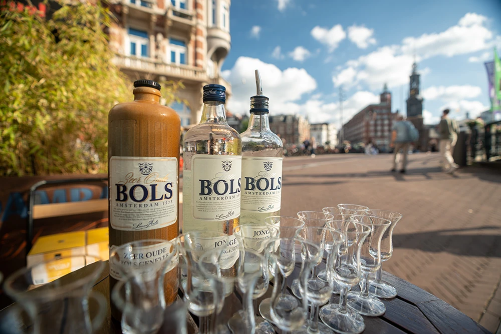 Three bottles of Bols Amsterdam genever and several empty tasting glasses on a wooden table outside in a sunny urban setting.