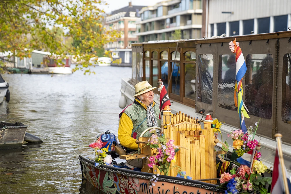 Man playing a wooden barrel organ on a flower-adorned boat in a canal with buildings and trees in the background.
