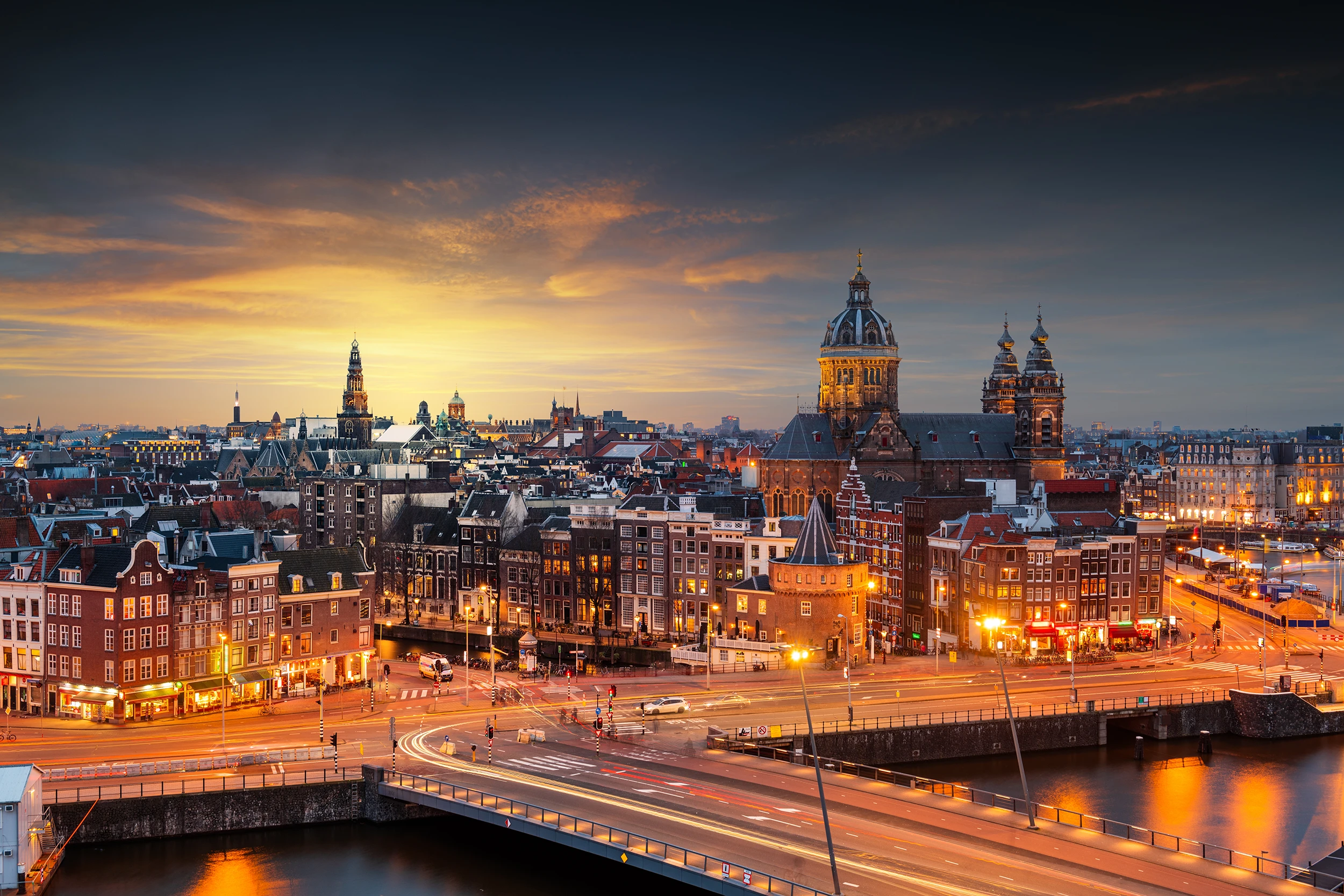 Evening cityscape of Amsterdam with illuminated historic buildings, canals, and a sunset sky.