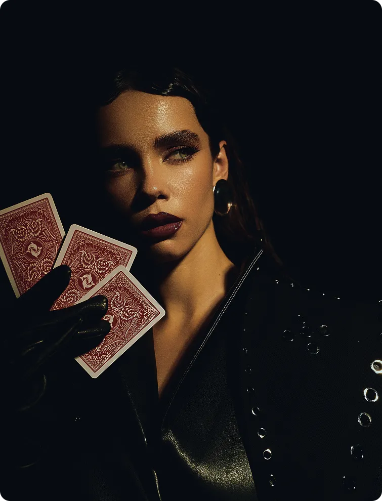 Woman wearing dark clothing and gloves holding three playing cards with red backs against a black background.