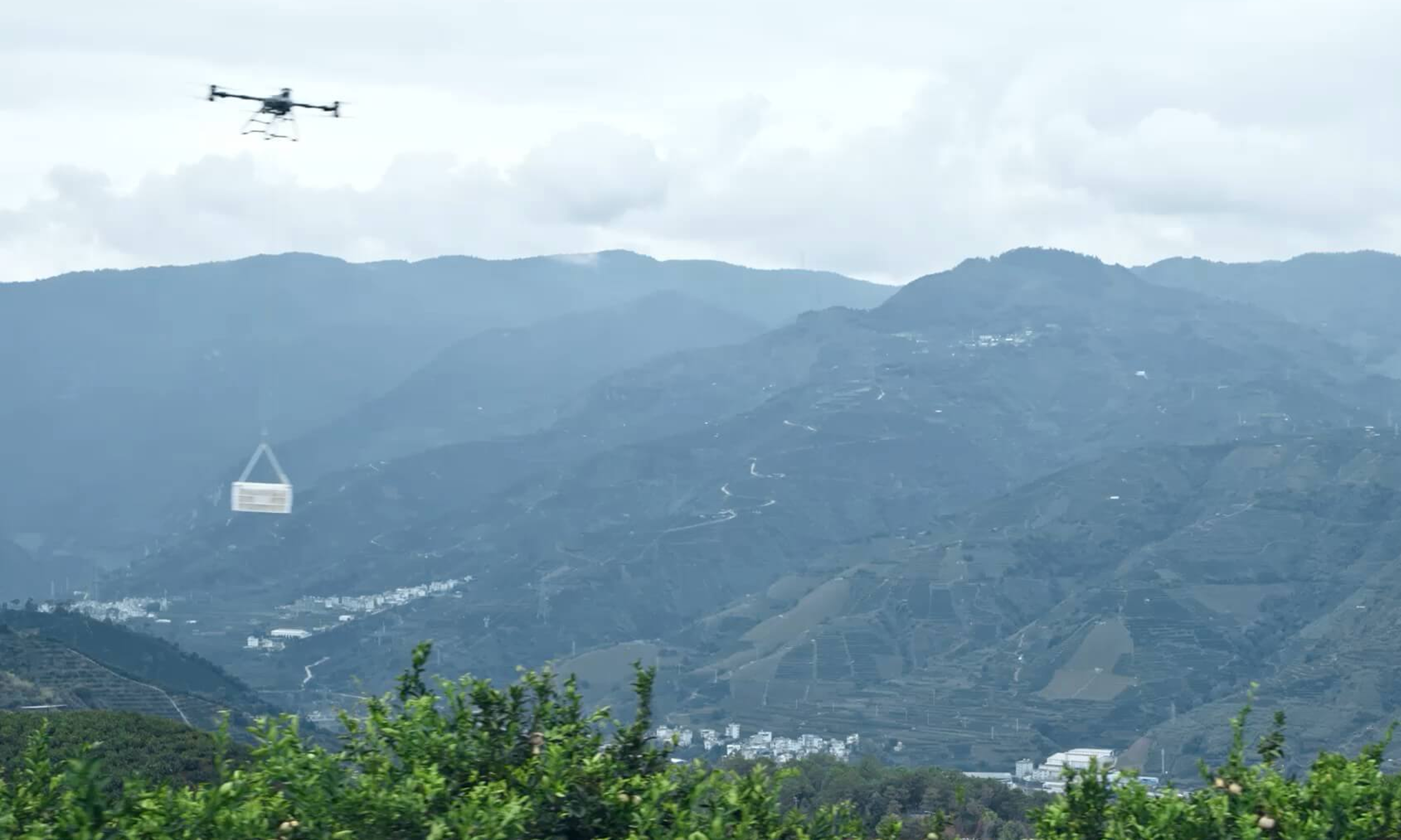 Drone flying in a mountainous landscape, carrying a suspended package below it.
