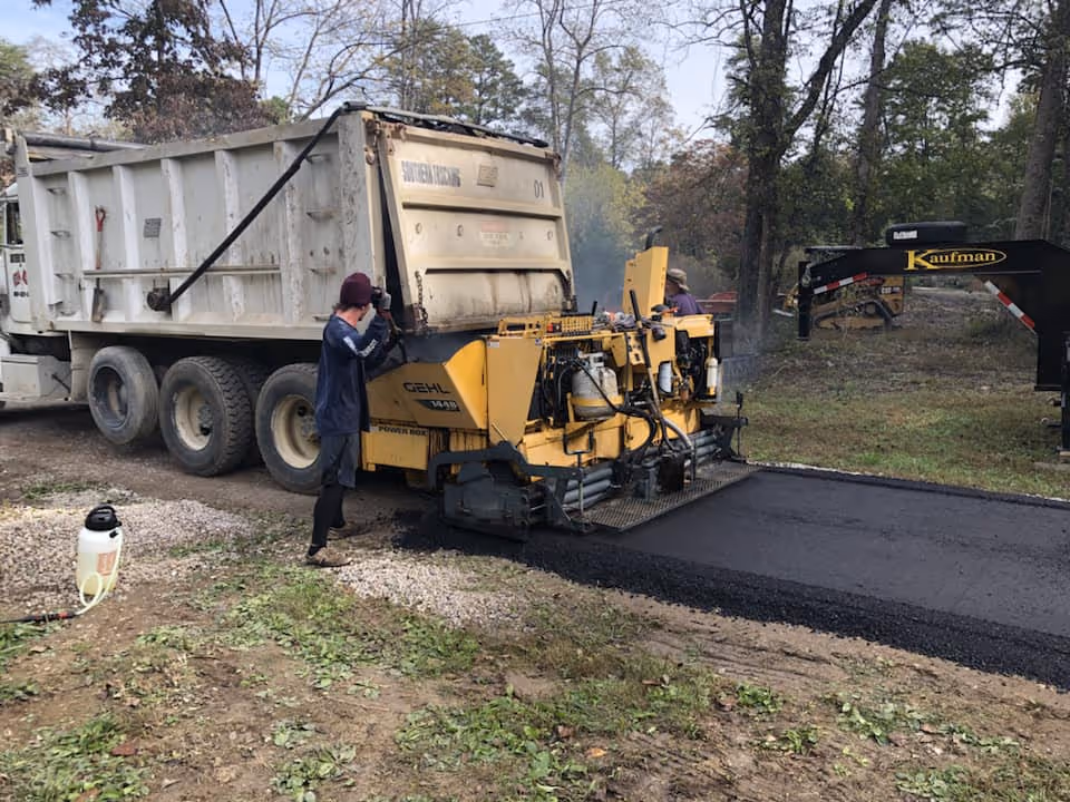 Construction workers operating a yellow asphalt paving machine laying fresh black asphalt beside a white dump truck in an outdoor, wooded area.