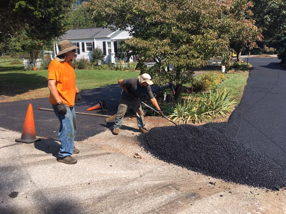 Two workers repaving a driveway with fresh black asphalt near a white house and green yard.