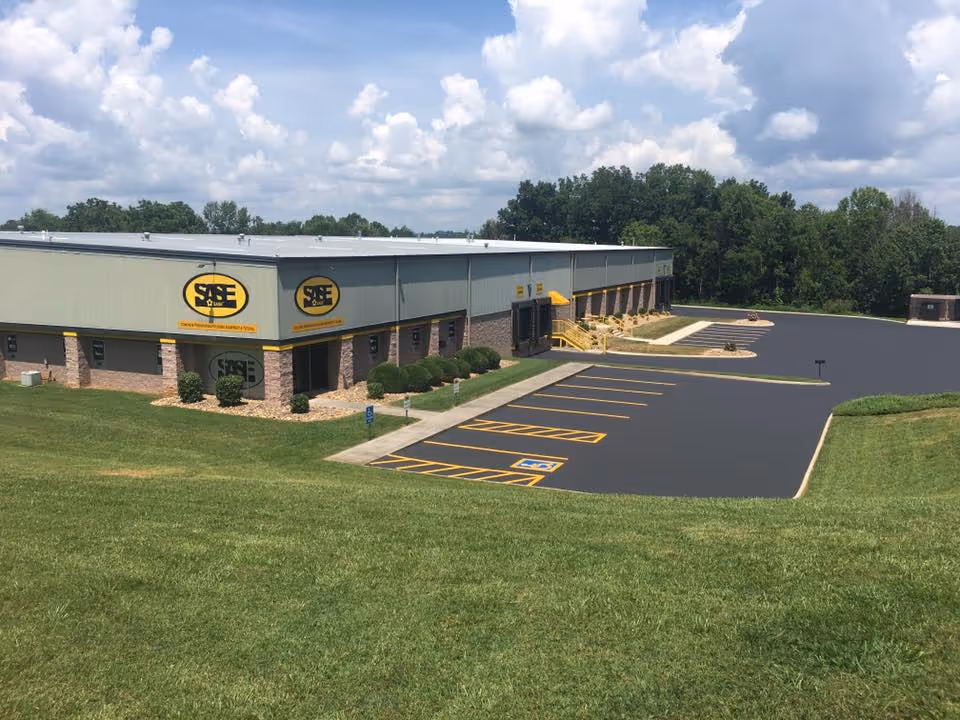 Industrial building with SAE International logos, surrounded by green grass and a parking lot with marked handicapped spaces under a cloudy sky.