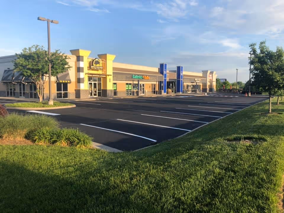 Empty parking lot in front of a shopping plaza with clear skies and green grass surrounding it.