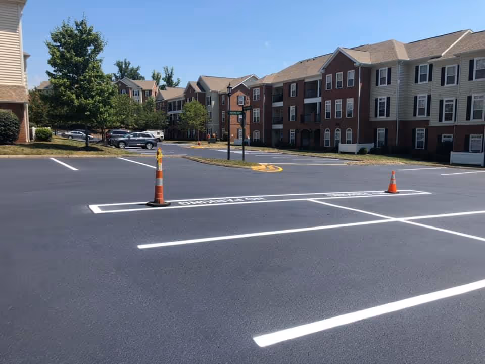 Empty parking lot with newly painted white lines and two orange traffic cones, surrounded by multi-story residential buildings and green trees under a clear blue sky.