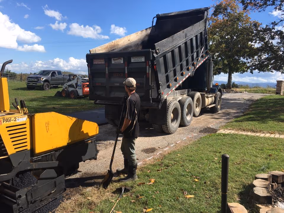 Man holding a shovel standing next to a yellow paving machine and a black dump truck with raised bed on a rural driveway.