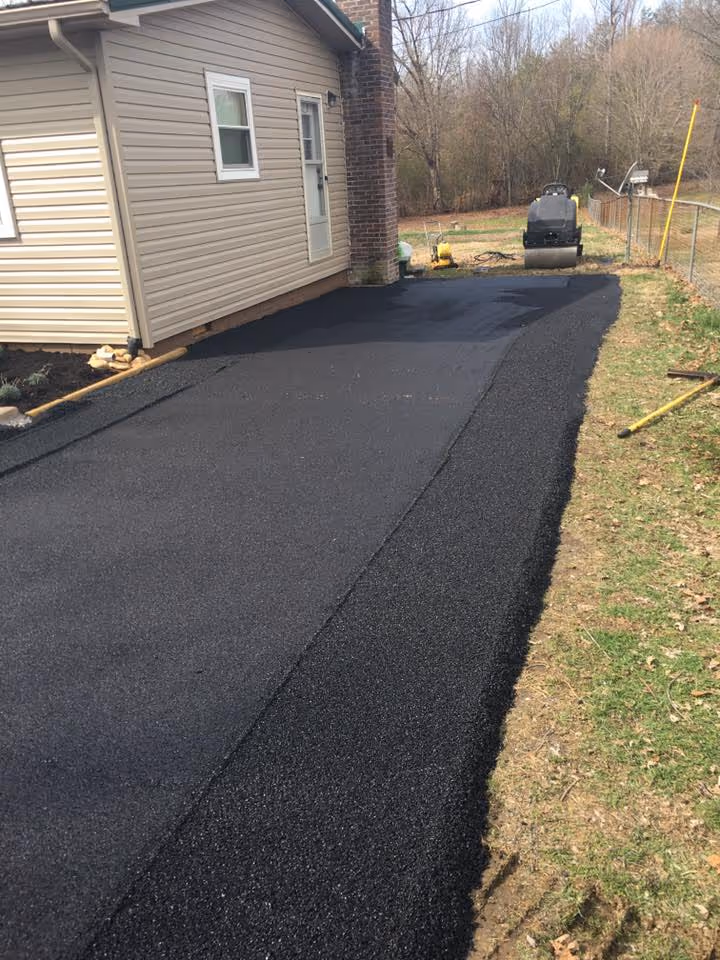Freshly paved black asphalt driveway next to a beige house with a brick chimney, bordered by grass and a chain-link fence.