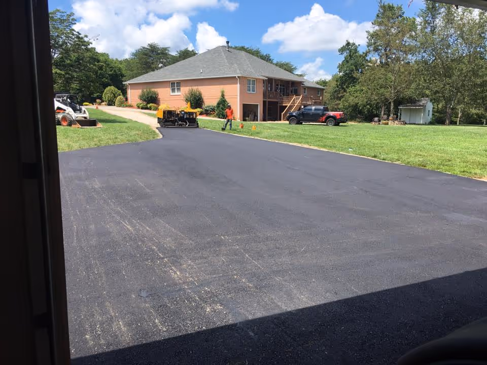 Freshly paved black asphalt driveway leading to a two-story brick house with a gray roof and surrounding green lawn on a partly cloudy day.