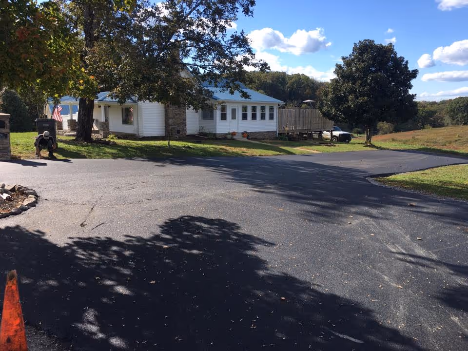 A newly paved driveway in front of a white house with a blue roof, surrounded by large trees under a partly cloudy sky.