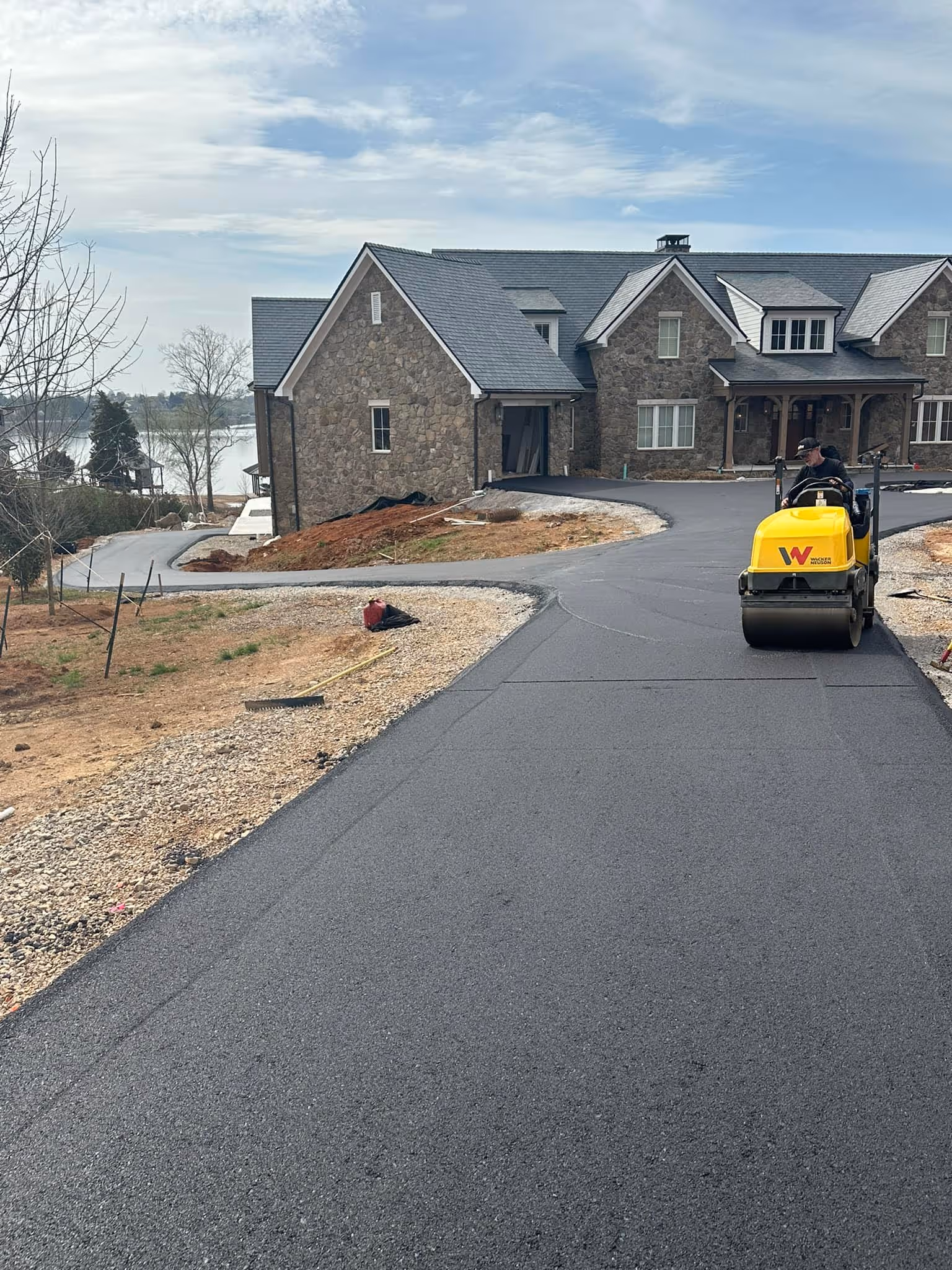 Newly paved asphalt driveway in front of a large stone house with a man operating a yellow road roller machine.
