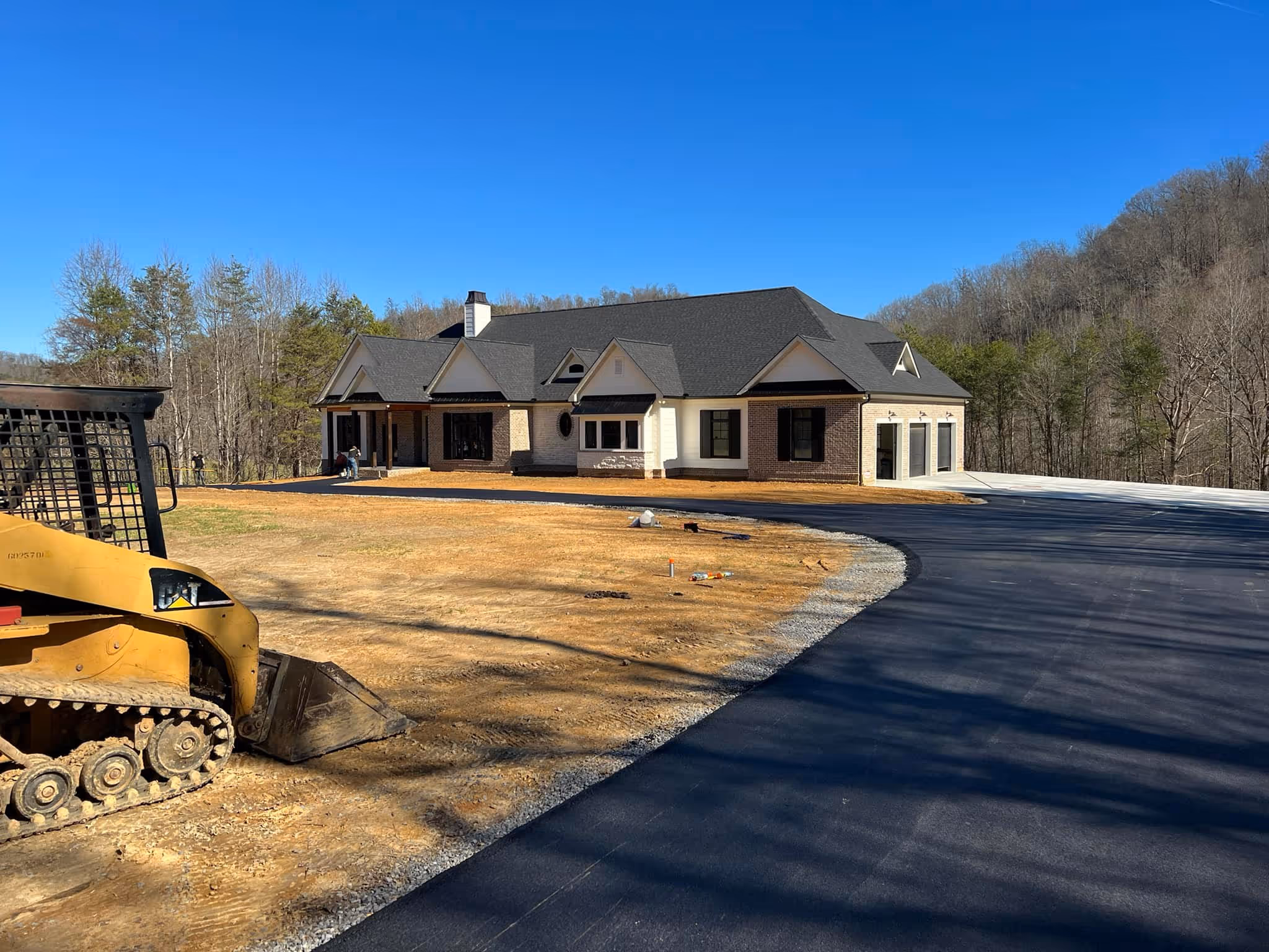 Large modern house with a dark roof, multiple gables, and a spacious paved driveway under clear blue sky.