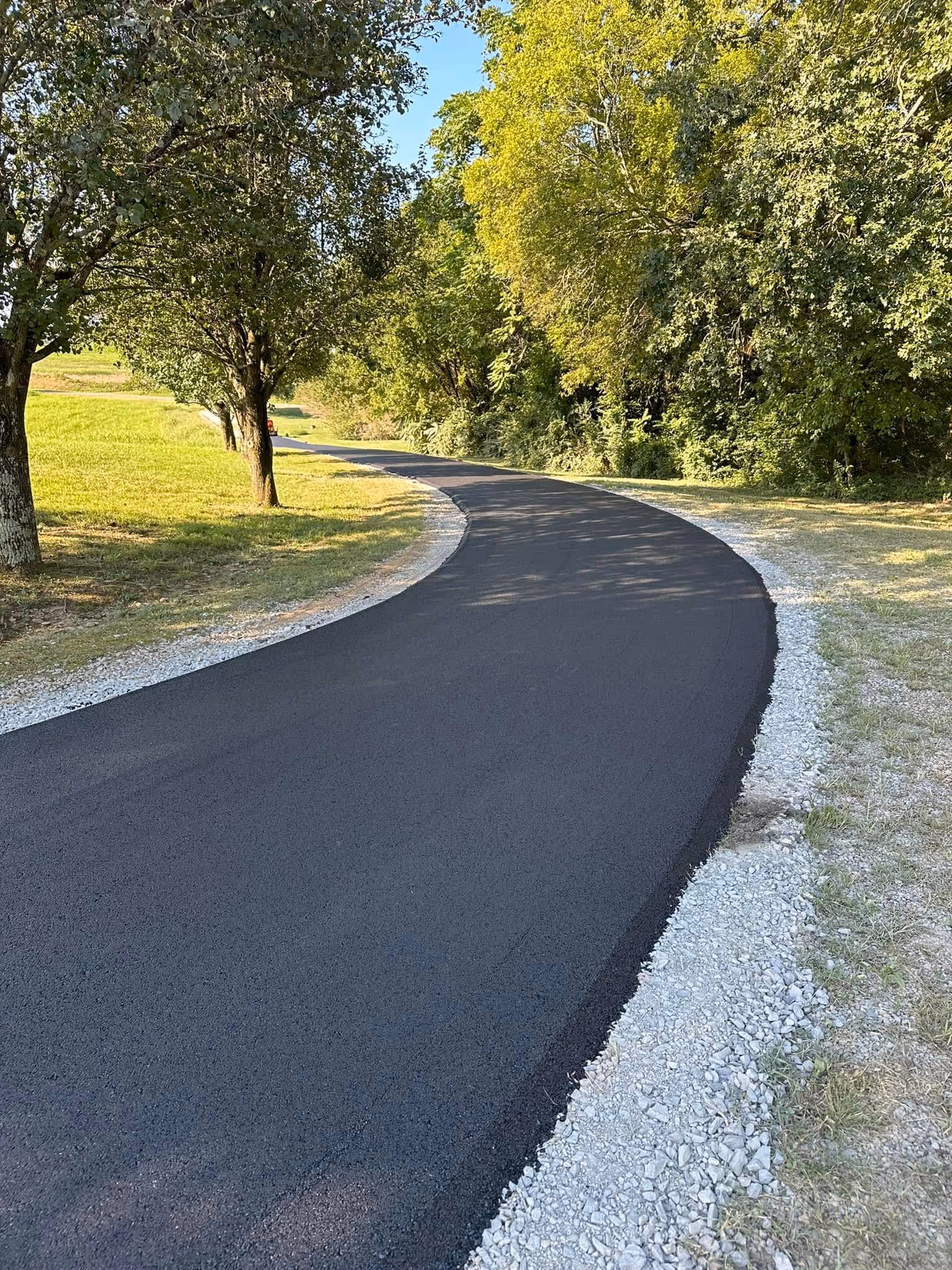 Newly paved winding asphalt path bordered by gravel, surrounded by green trees and grass under a clear blue sky.