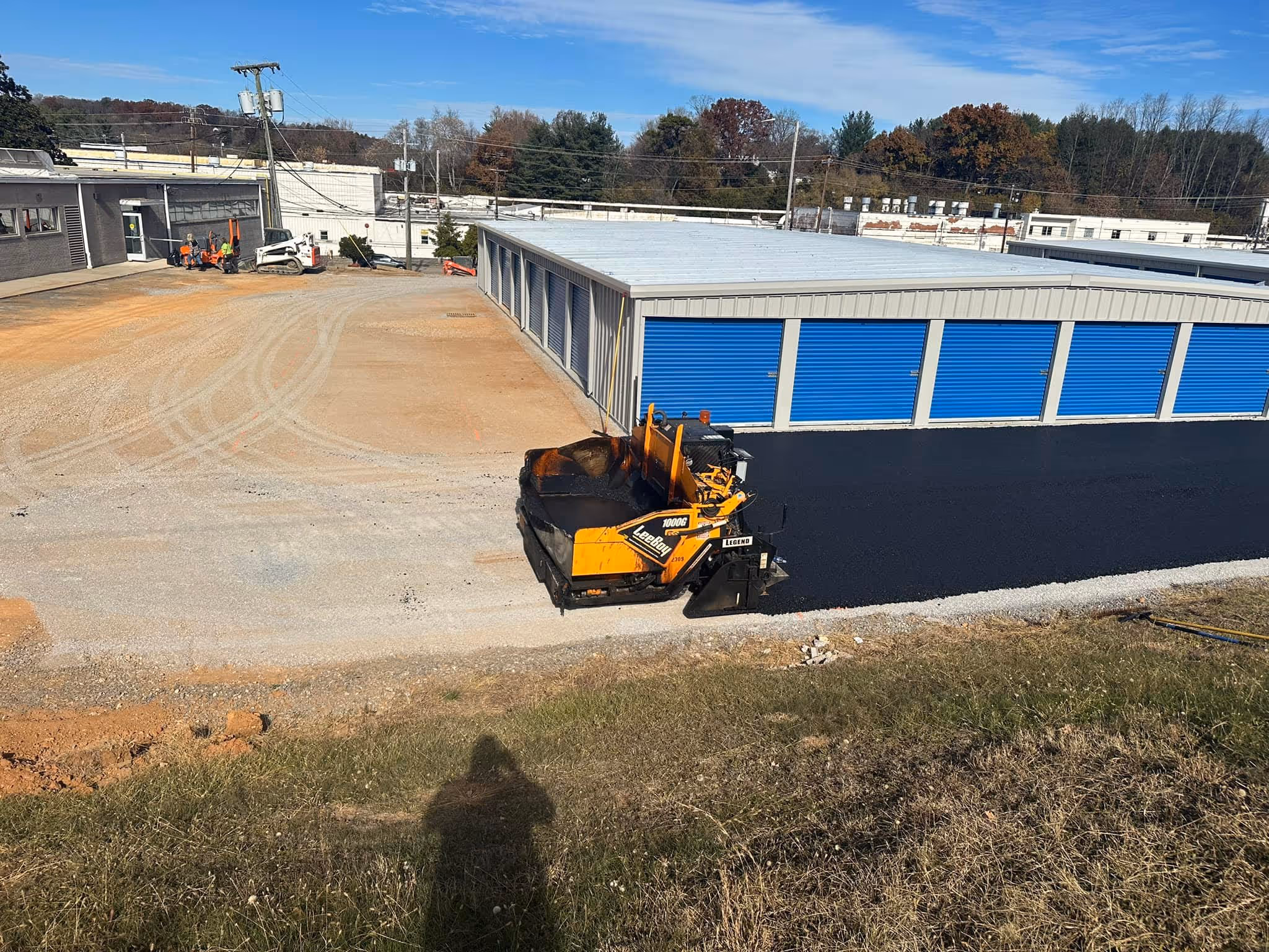 Construction scene with a yellow asphalt paver laying black asphalt in front of a storage facility with blue doors under a clear sky.