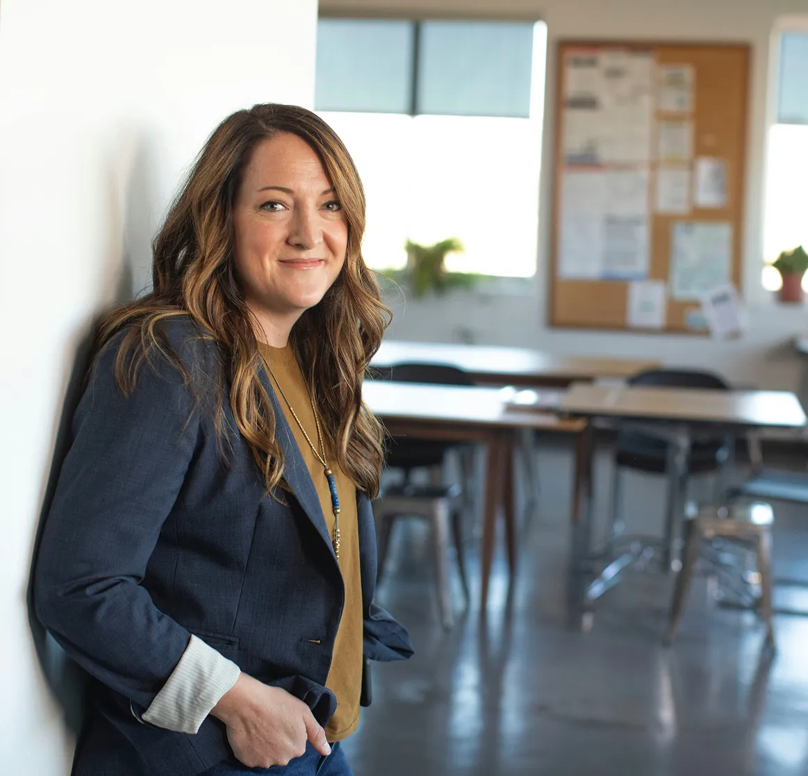 Smiling woman with long wavy hair wearing a navy blazer and mustard shirt leaning against a white wall in an office setting.