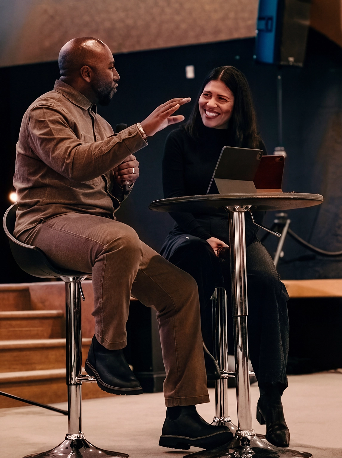 A man and woman sitting on high stools at a round table engaged in a conversation, the man gesturing with his hand and the woman smiling.