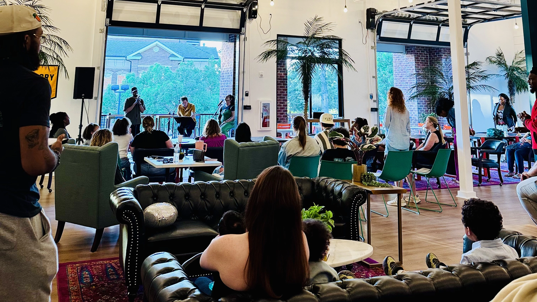Group of people seated in a modern indoor space watching a panel of three speakers near large windows with trees outside.