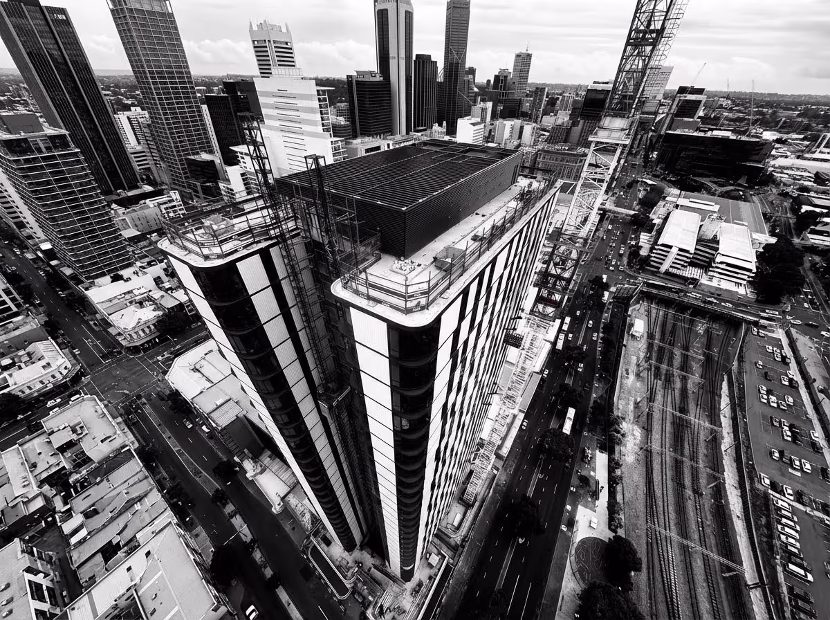Aerial view of a modern cityscape featuring a tall building under construction with cranes and surrounding roads and railway tracks.