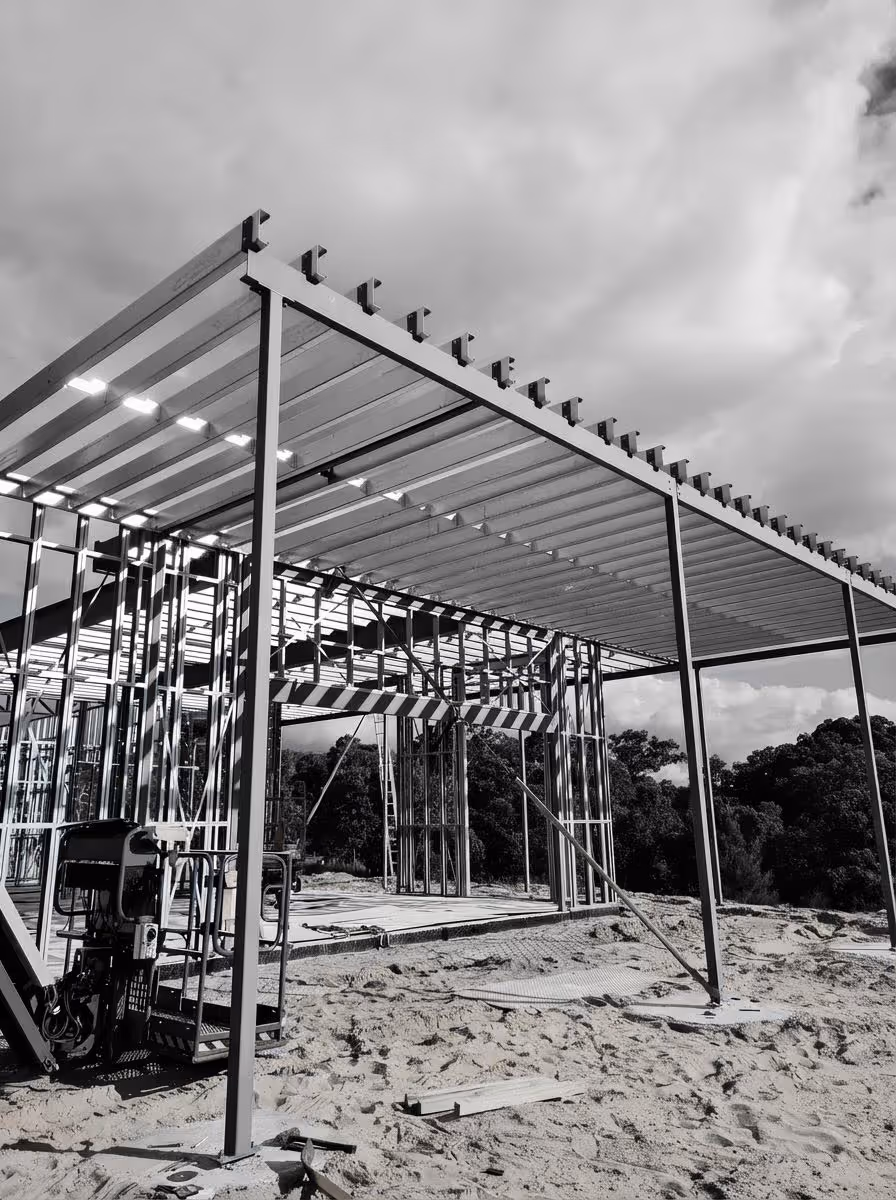 Steel frame structure under construction on a sandy site with trees in the background and a cloudy sky.