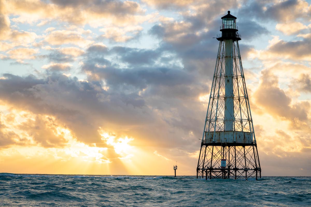 Metal lighthouse structure in the ocean during a cloudy sunrise with sun rays over the horizon.