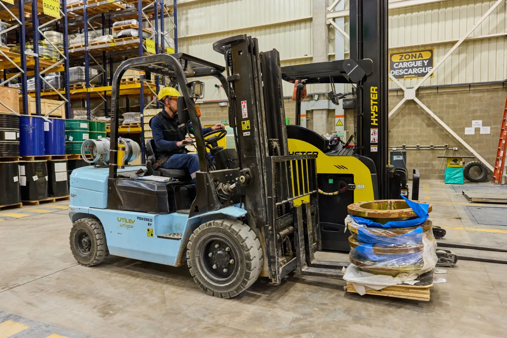 Warehouse worker wearing a yellow helmet operating a forklift lifting wrapped metal parts on a pallet.