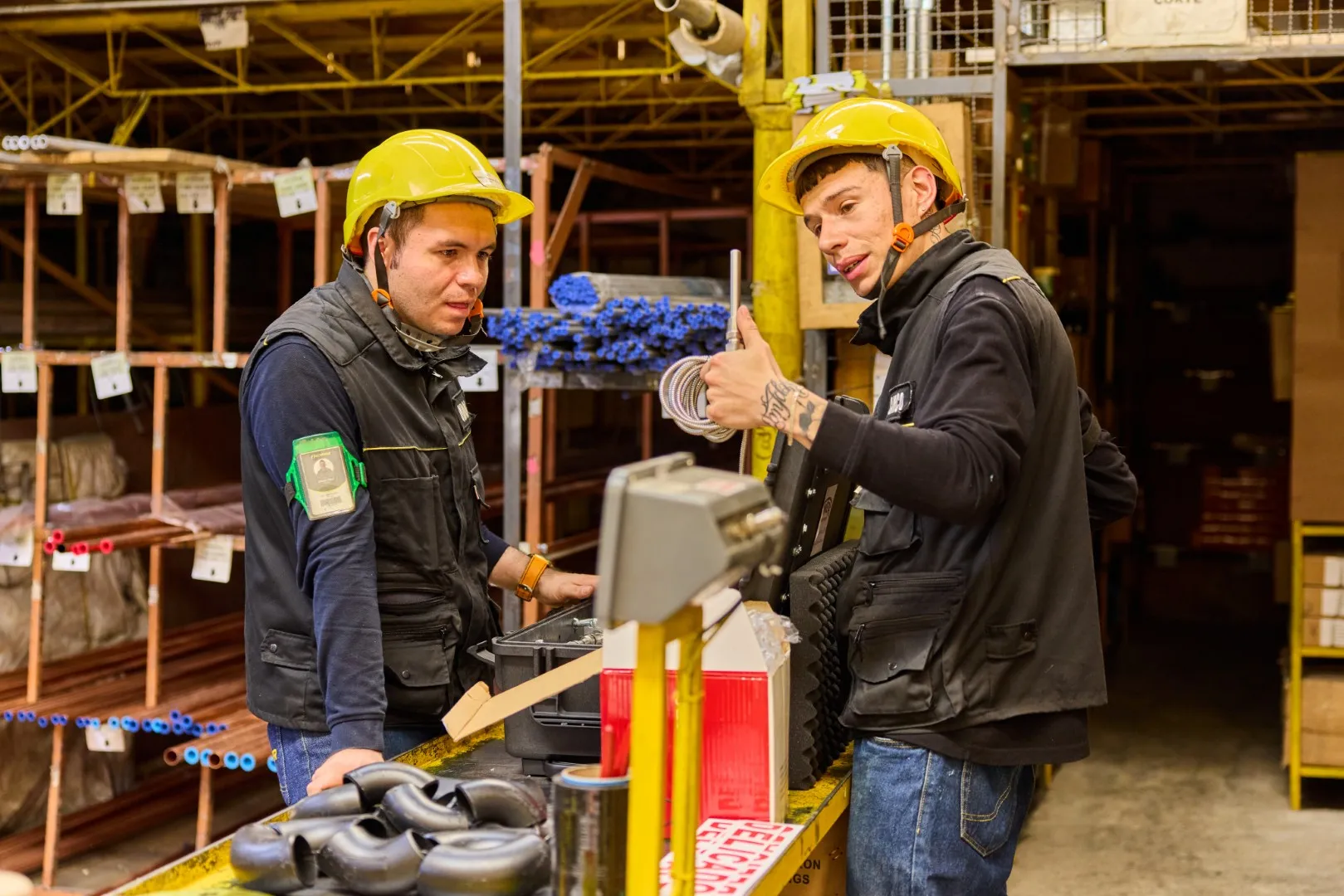 Two warehouse workers wearing yellow hard hats discuss tools and supplies on a workbench.