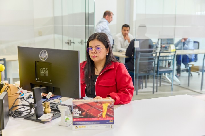 Woman with glasses in a red jacket working at a computer in an office with people in the background.
