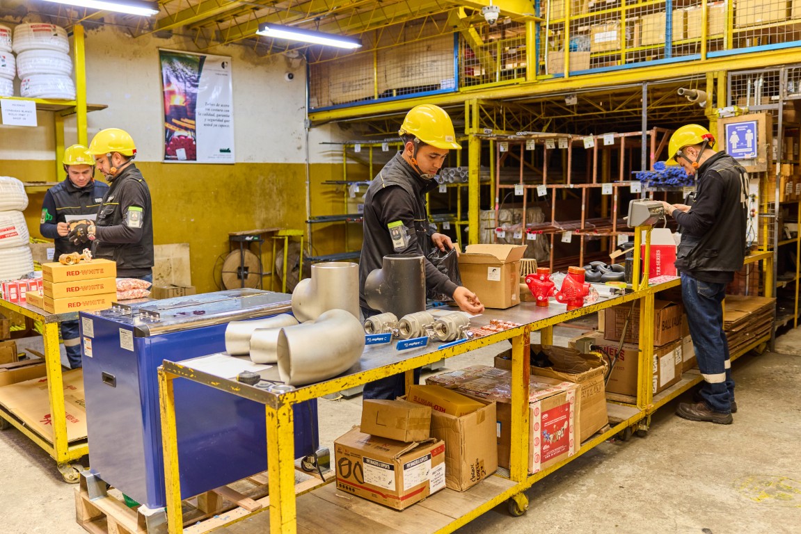 Workers in yellow helmets packaging industrial parts and equipment on a long yellow table in a storage area.