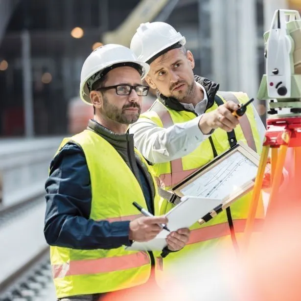 Two engineers in hard hats and safety vests reviewing plans and surveying on a railway construction site.
