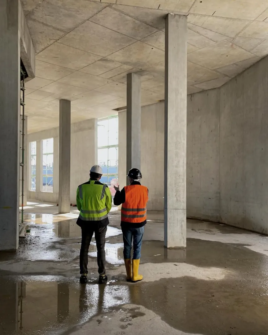 Two construction workers wearing safety vests and helmets inspecting a wet concrete floor indoors.