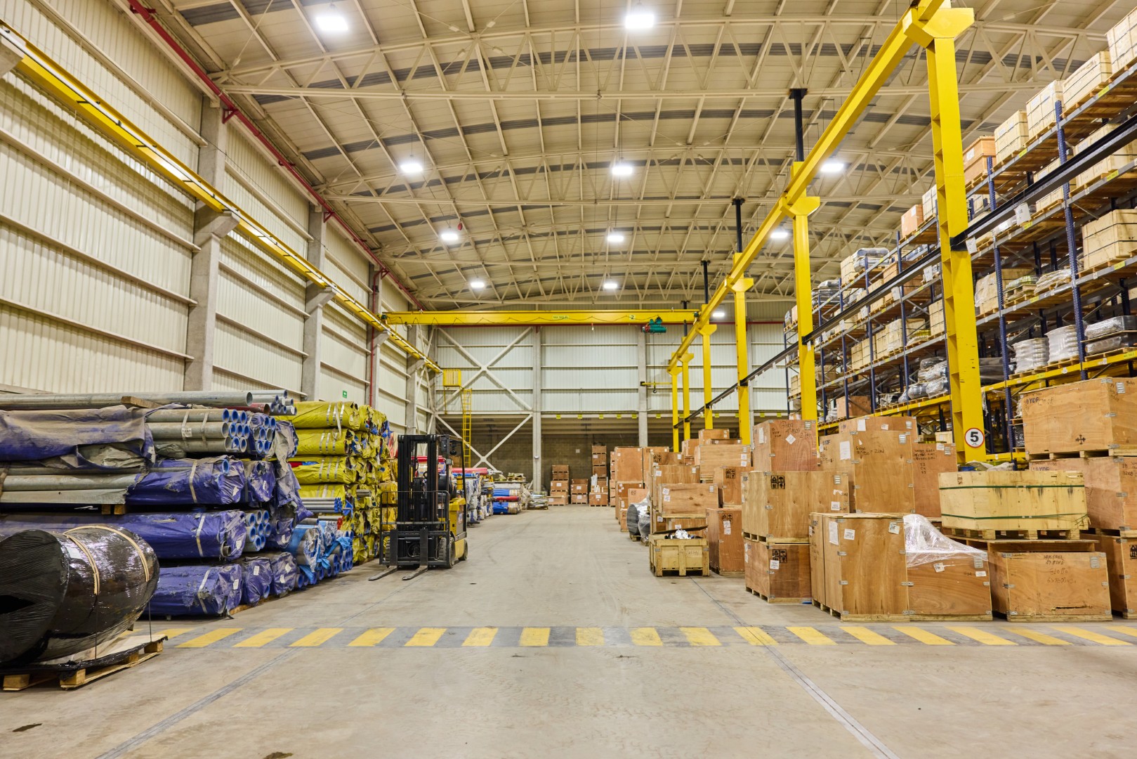 Spacious warehouse with stacked wooden crates, wrapped pipes, and a forklift on a wide aisle.