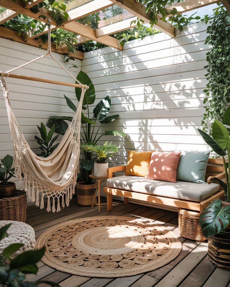 Sunny enclosed patio deck with macrame hammock chair, wood bench with colorful cushions, jute rug, and an abundance of tropical and monstera potted plants under a pergola.