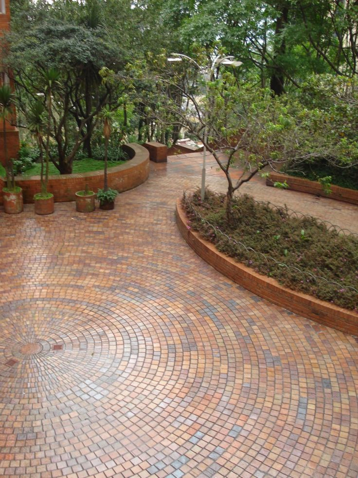 Aerial view of a large courtyard with circular patterned multi-tone brick pavers, curved raised garden beds with trees and shrubs, and lush tree canopy overhead.