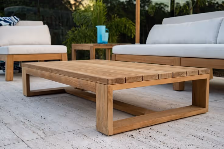Close-up of a solid teak wood outdoor coffee table with slatted top on a white stone patio, flanked by white cushion teak sofas with tropical plants in background.