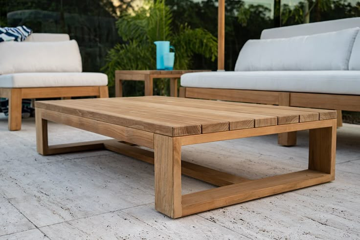 Close-up of a solid teak wood outdoor coffee table with slatted top on a white stone patio, flanked by white cushion teak sofas with tropical plants in background.