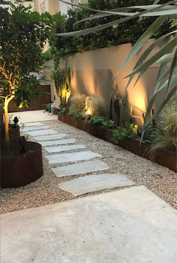 Narrow side yard pathway with irregular stepping stones through decomposed granite gravel, corten steel raised planters with agave and grasses, and warm uplighting on stucco wall at dusk.