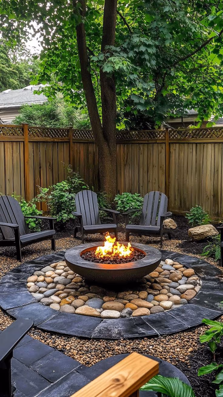 Circular backyard fire pit area with slate stepping stone base, decorative river rock ring, round concrete fire bowl with flames, dark Adirondack chairs, and mature tree overhead.