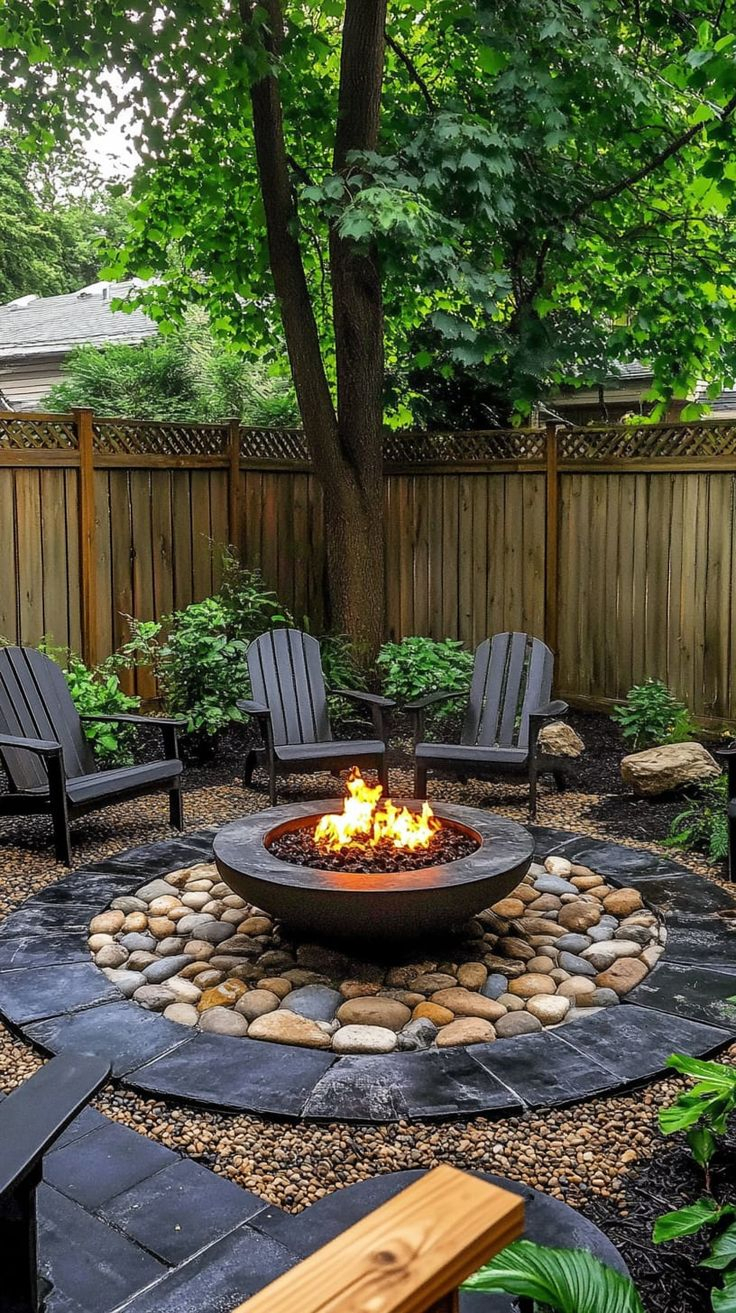 Circular backyard fire pit area with slate stepping stone base, decorative river rock ring, round concrete fire bowl with flames, dark Adirondack chairs, and mature tree overhead.