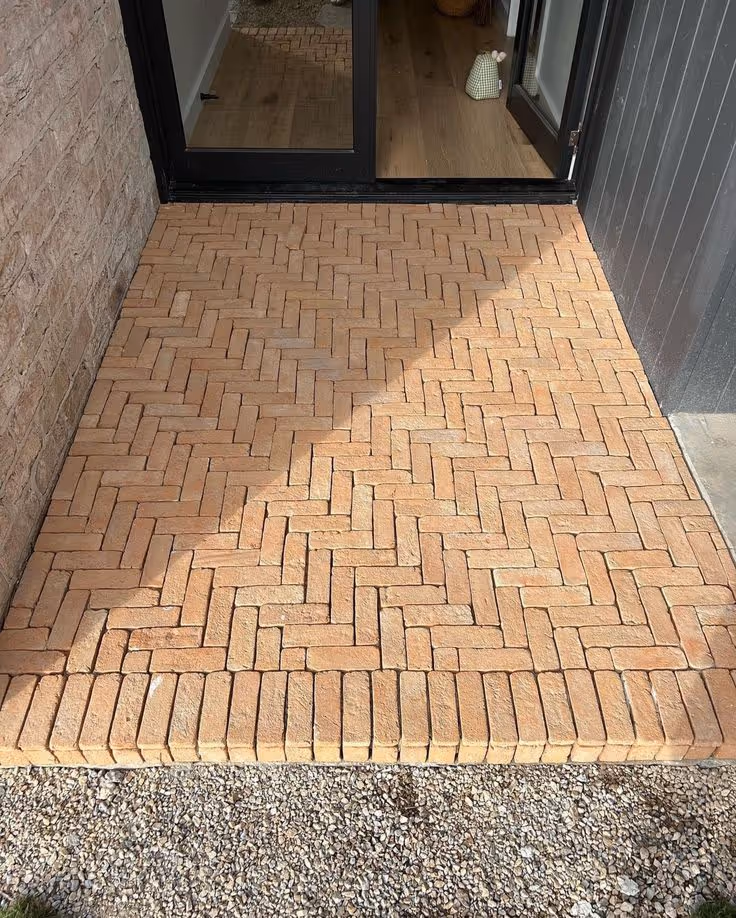 Small terracotta herringbone-patterned brick paver patio at the base of sliding glass doors, with gravel border and adjacent brick wall.