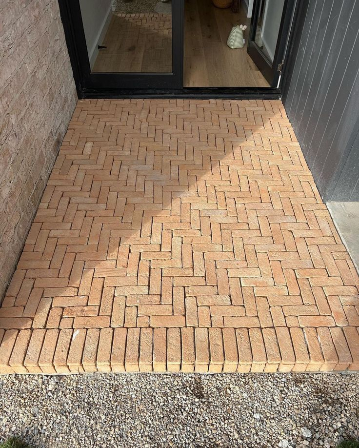 Small terracotta herringbone-patterned brick paver patio at the base of sliding glass doors, with gravel border and adjacent brick wall.