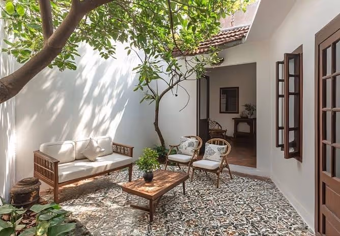 White stucco enclosed Mediterranean courtyard with decorative encaustic tile floor, mature shade tree, teak sofa and chairs with white cushions, and interior visible through open doorway.