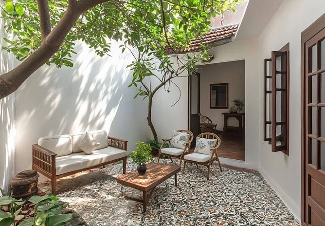 White stucco enclosed Mediterranean courtyard with decorative encaustic tile floor, mature shade tree, teak sofa and chairs with white cushions, and interior visible through open doorway.