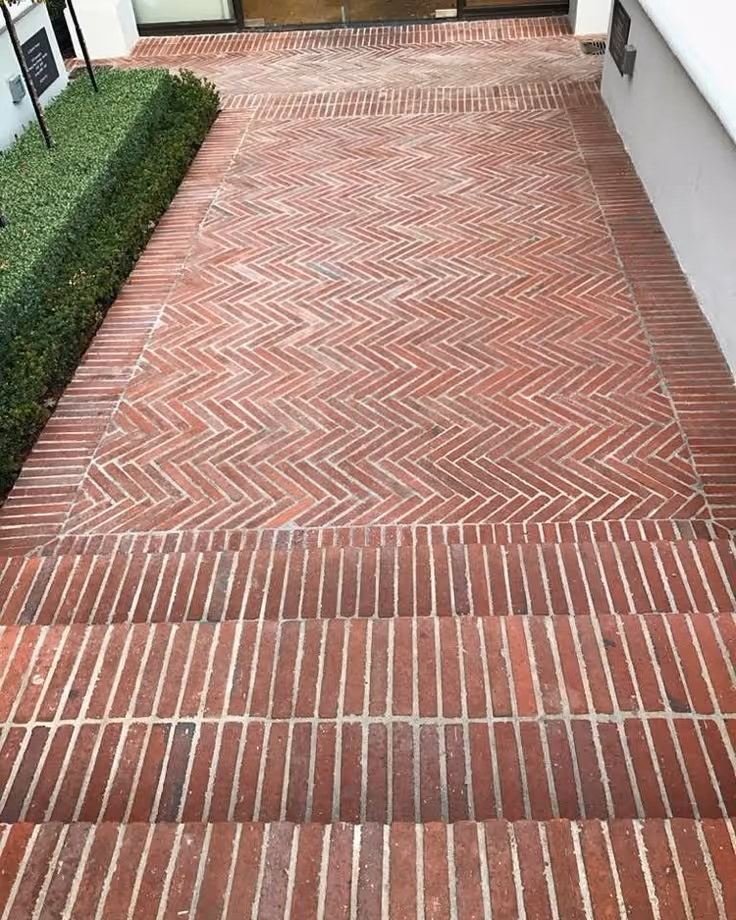 Classic red herringbone-patterned brick front entry paving with contrasting straight border course, boxwood hedge to the side, adjacent to white exterior wall.